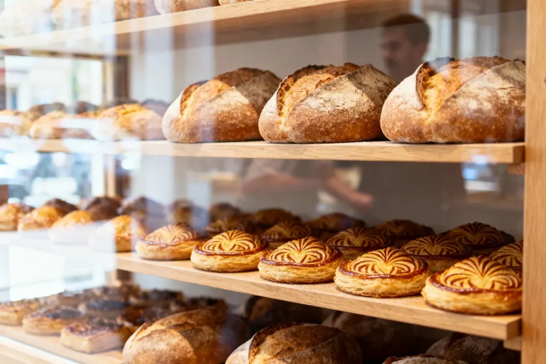 Étagères de boulangerie avec pains artisanaux et pâtisseries appétissantes, reflet d'un homme en arrière-plan.