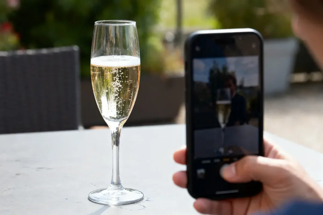 Un verre de champagne est photographié par une personne utilisant un smartphone, en extérieur.