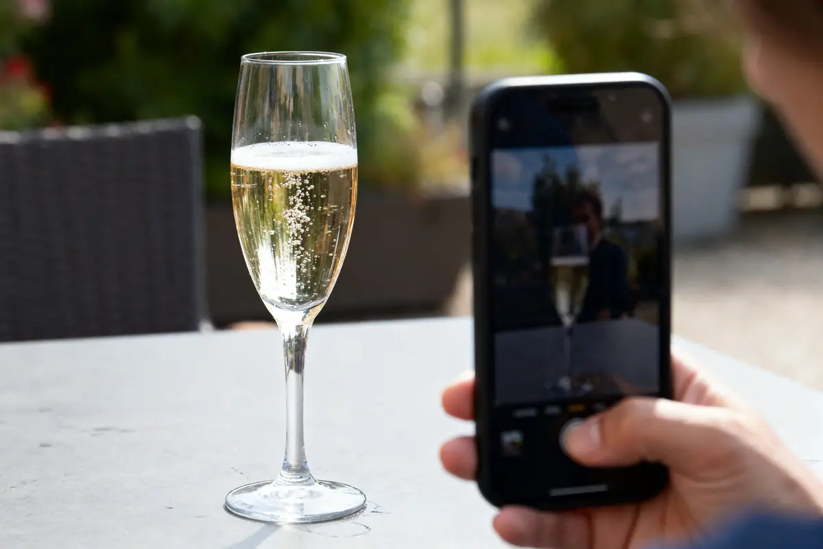 Un verre de champagne est photographié par une personne utilisant un smartphone, en extérieur.