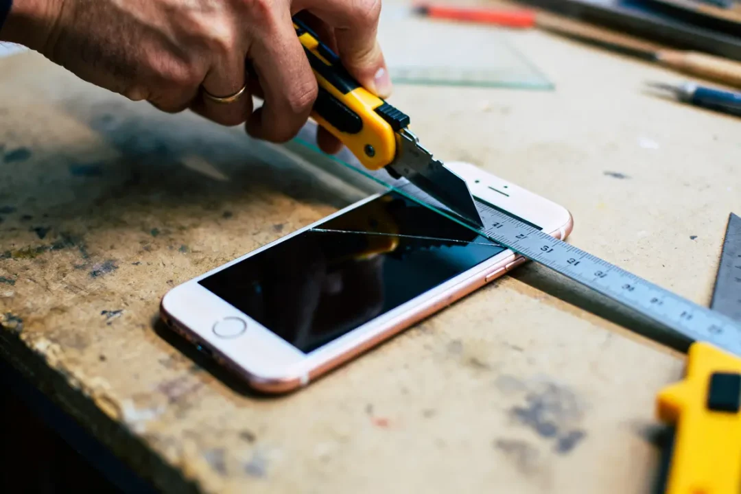 Main coupant verre au-dessus d'un smartphone avec cutter et règle métallique sur table atelier.