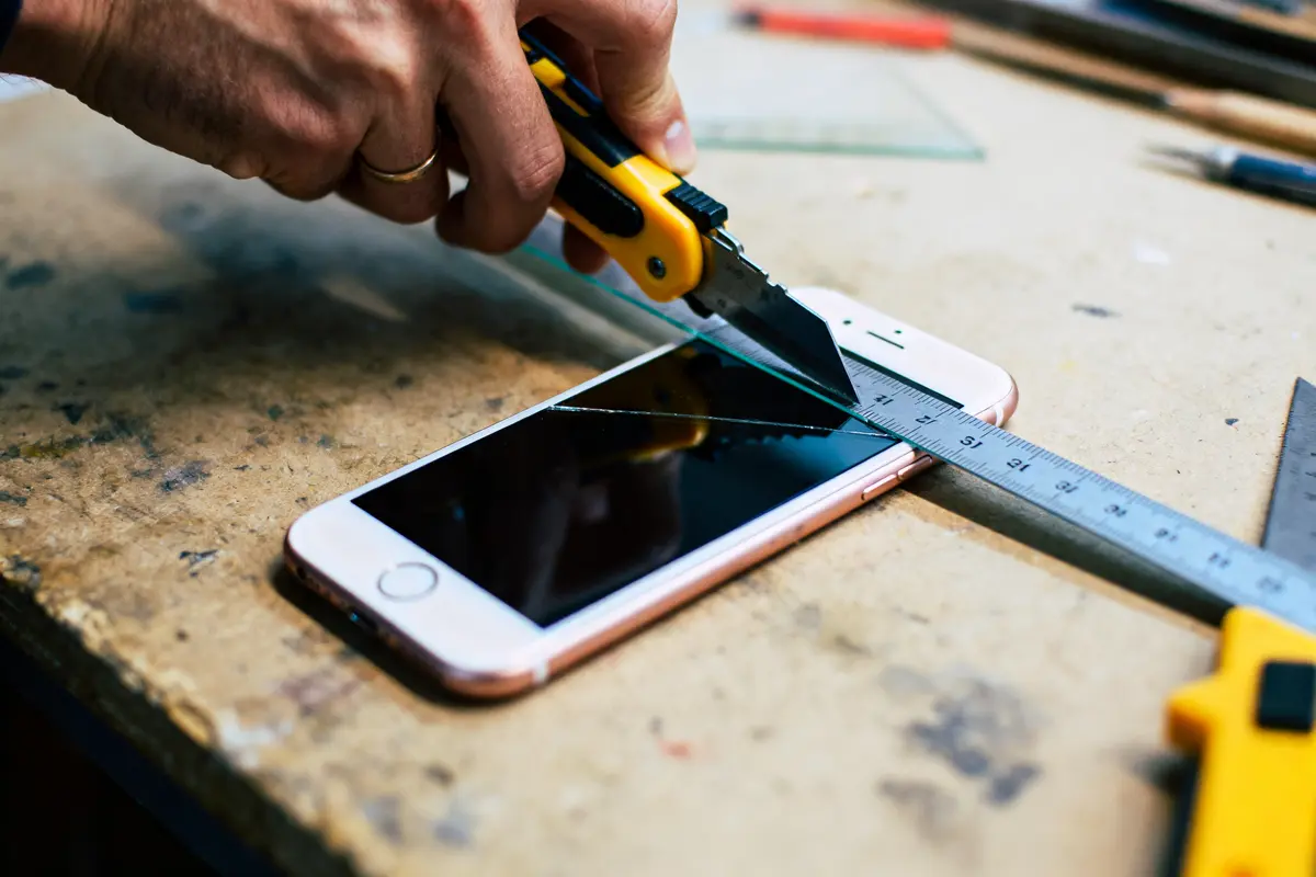 Main coupant verre au-dessus d'un smartphone avec cutter et règle métallique sur table atelier.
