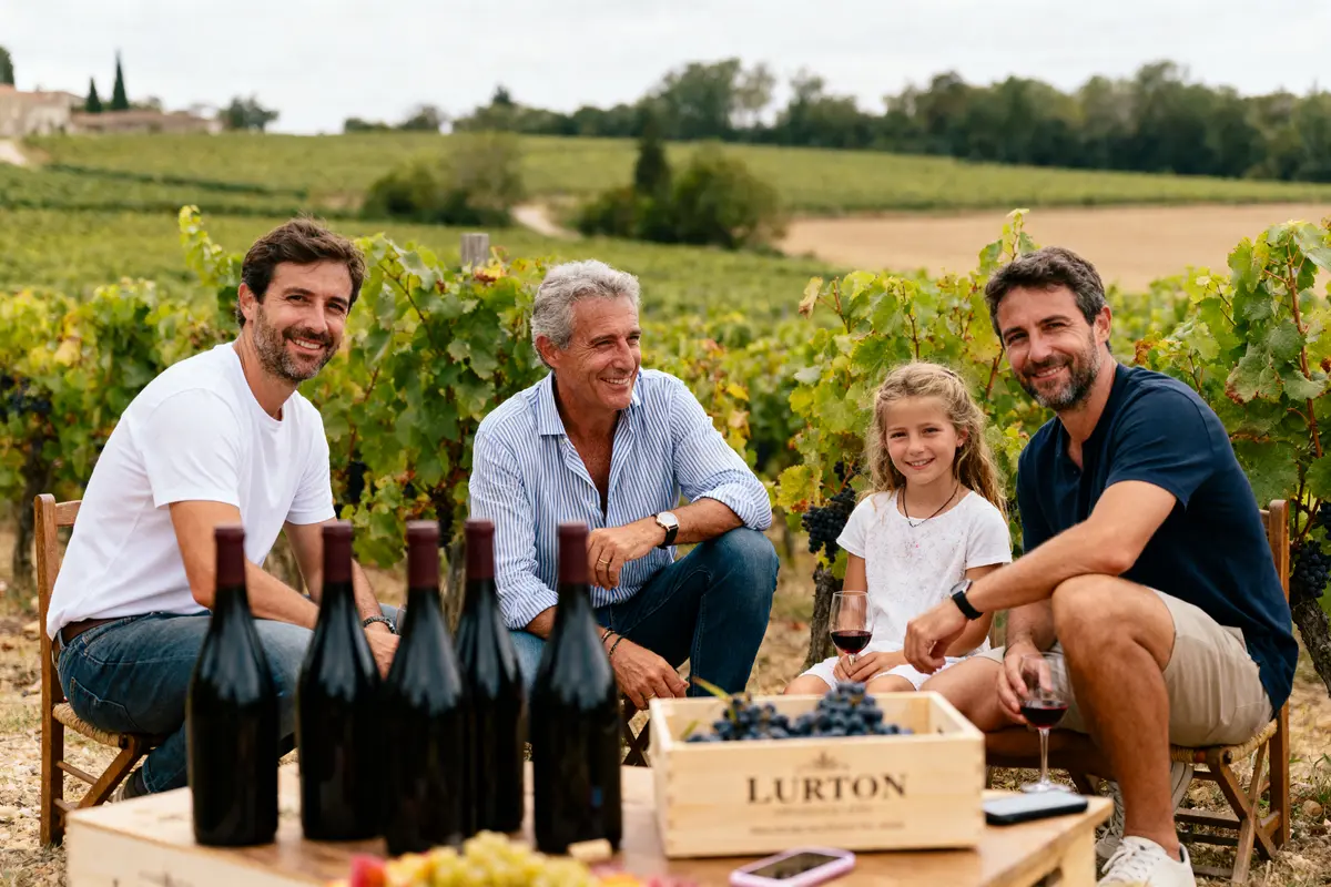 Personnes souriant dans un vignoble, assis autour d'une table avec des bouteilles et une caisse de raisins.