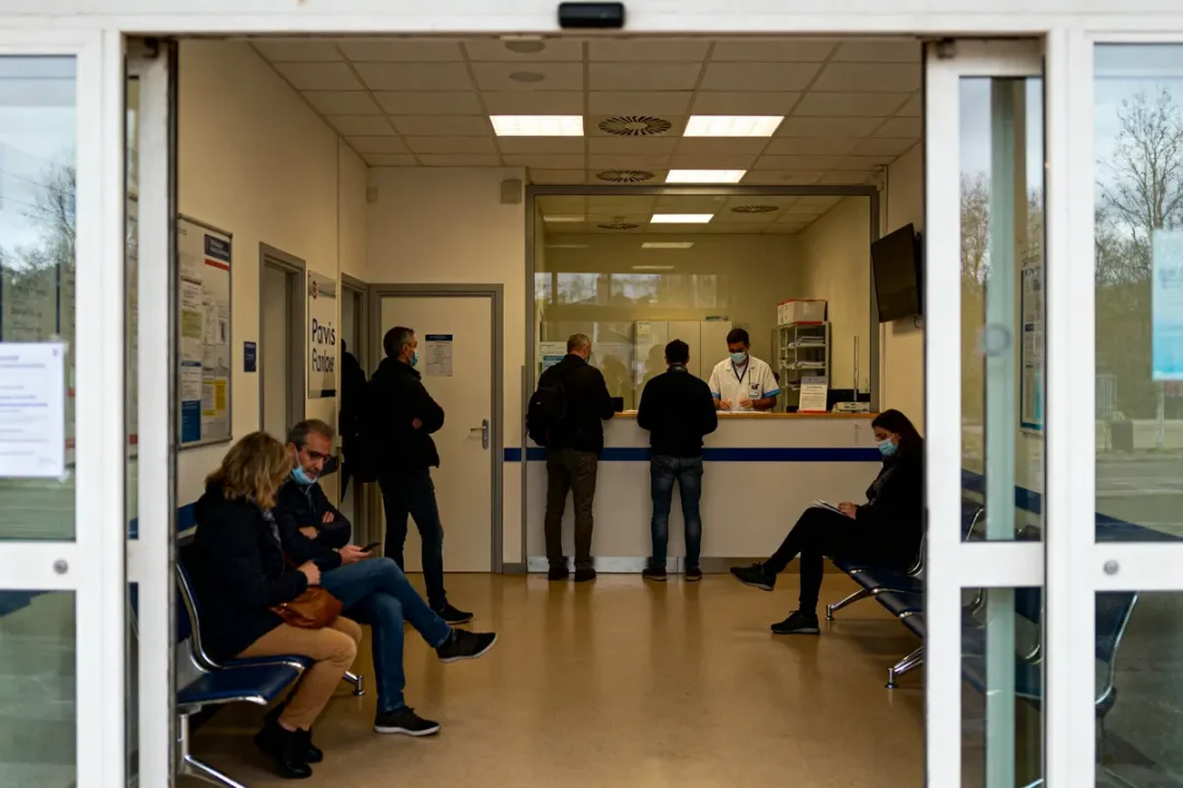 Salle d'attente d'un centre médical avec plusieurs personnes portant des masques et un réceptionniste derrière un comptoir.