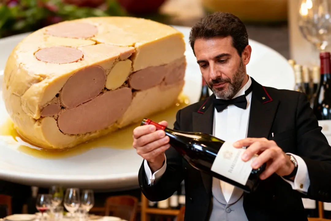 Un homme en smoking examine une bouteille de vin, avec du foie gras en arrière-plan.