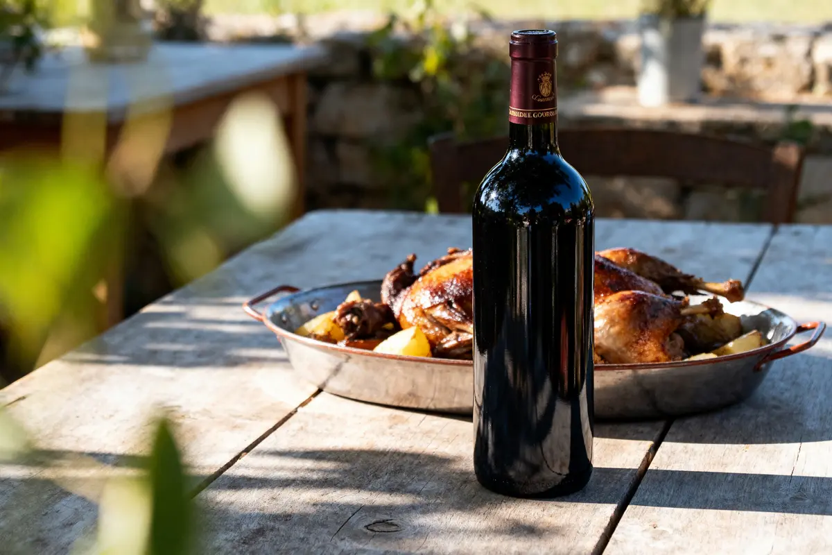 Bouteille de vin rouge devant un plat de poulet rôti avec légumes, sur table en bois extérieur.