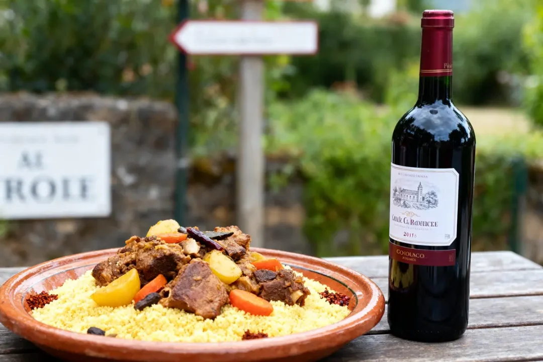 Plat de couscous garni de légumes et viande, accompagné d'une bouteille de vin rouge sur table extérieure.