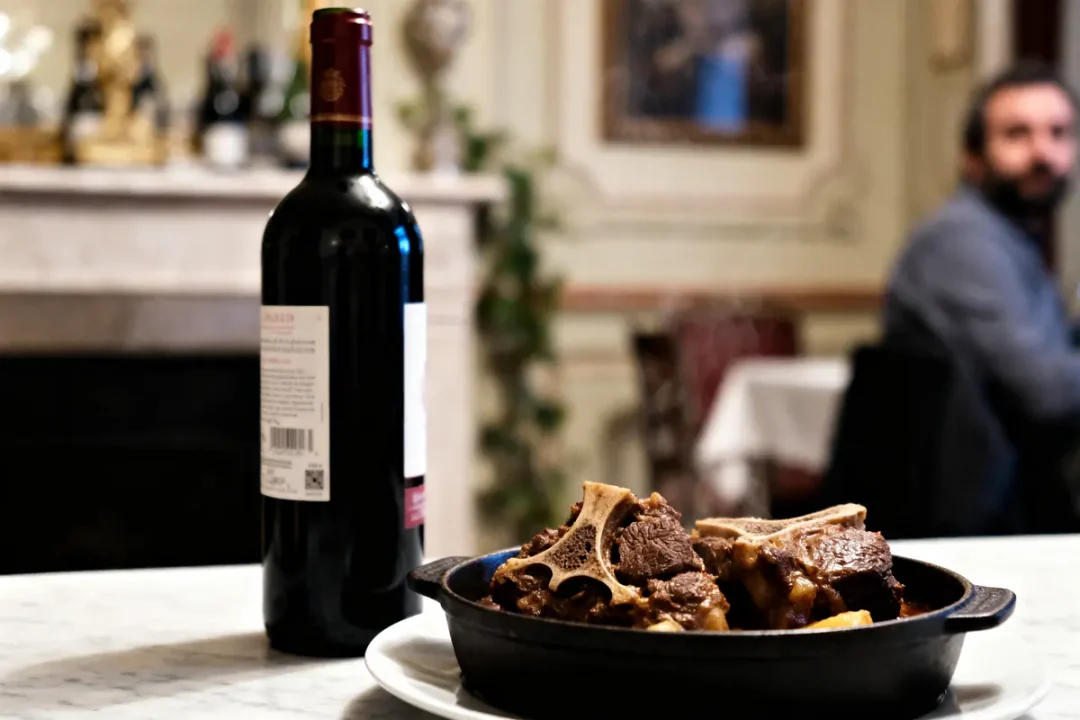 Bouteille de vin rouge et plat de viande braisée sur une table en marbre, homme assis en arrière-plan.