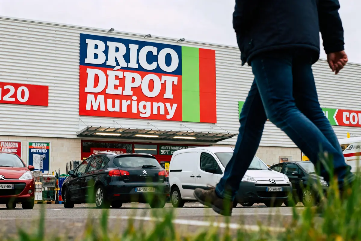 Façade d'un magasin Brico Dépôt avec parking, voitures, et une personne qui marche au premier plan.
