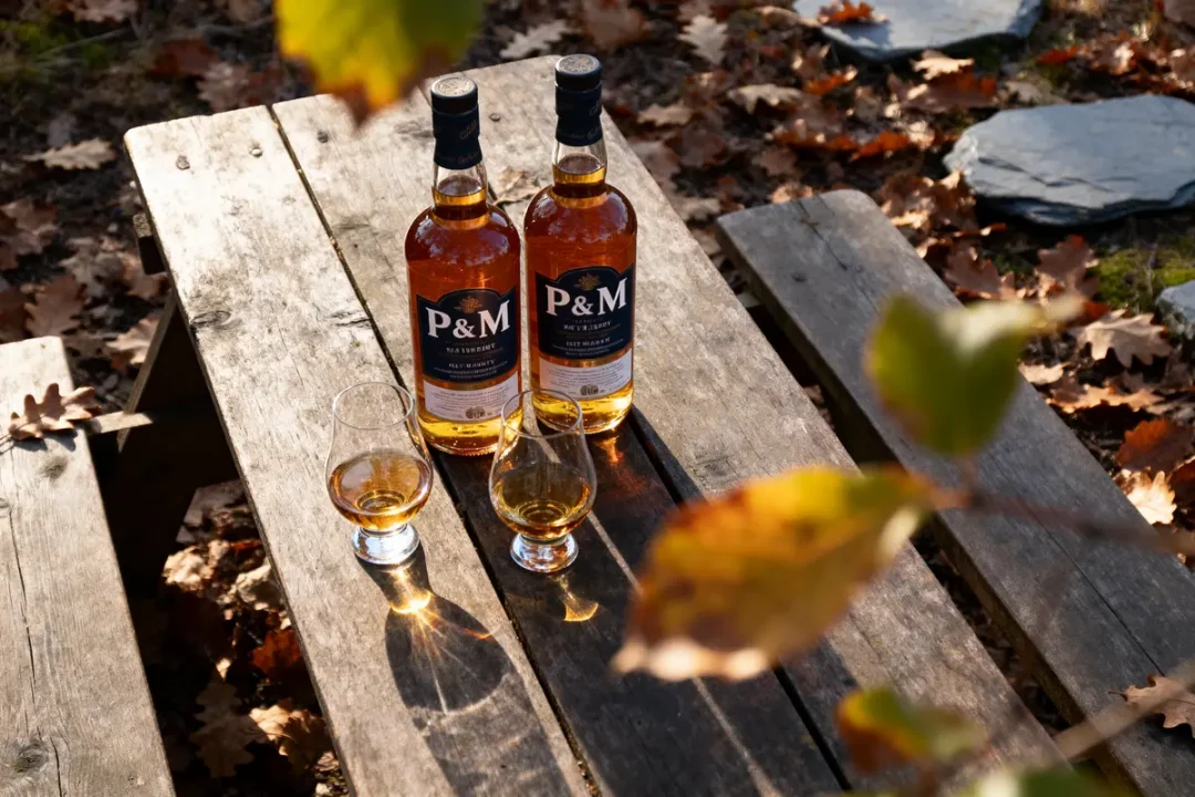 Deux bouteilles de whisky P&M et trois verres sur table en bois, entourés de feuilles d'automne.