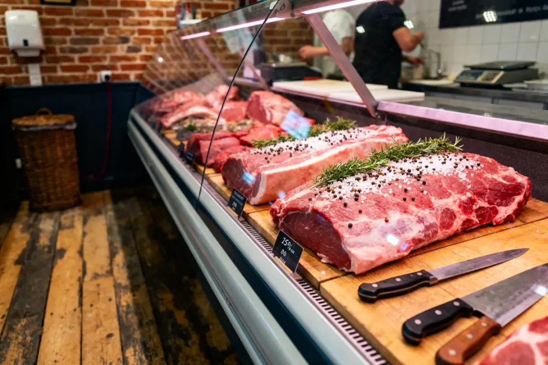 Viande crue en vitrine de boucherie, avec épices et couteaux à proximité. Décor de murs en briques.
