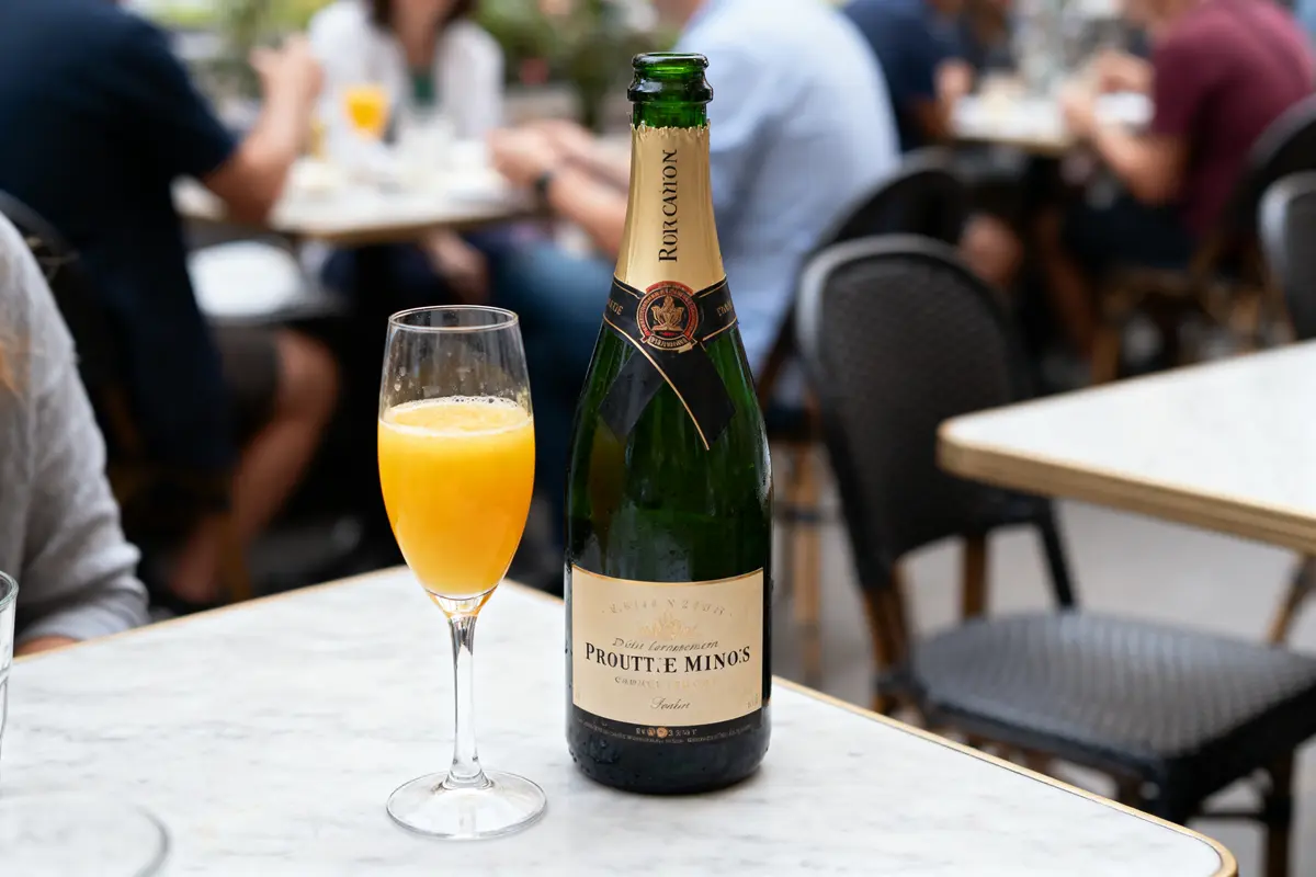 Bouteille de champagne et verre de cocktail orange sur table de café en extérieur, personnes en arrière-plan.