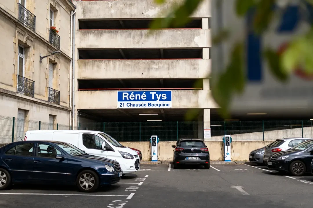 Parking avec voitures, bâtiment beige, panneau "Réné Tys", bornes de recharge. Adresse : 21 Chaussée Bocquaine.