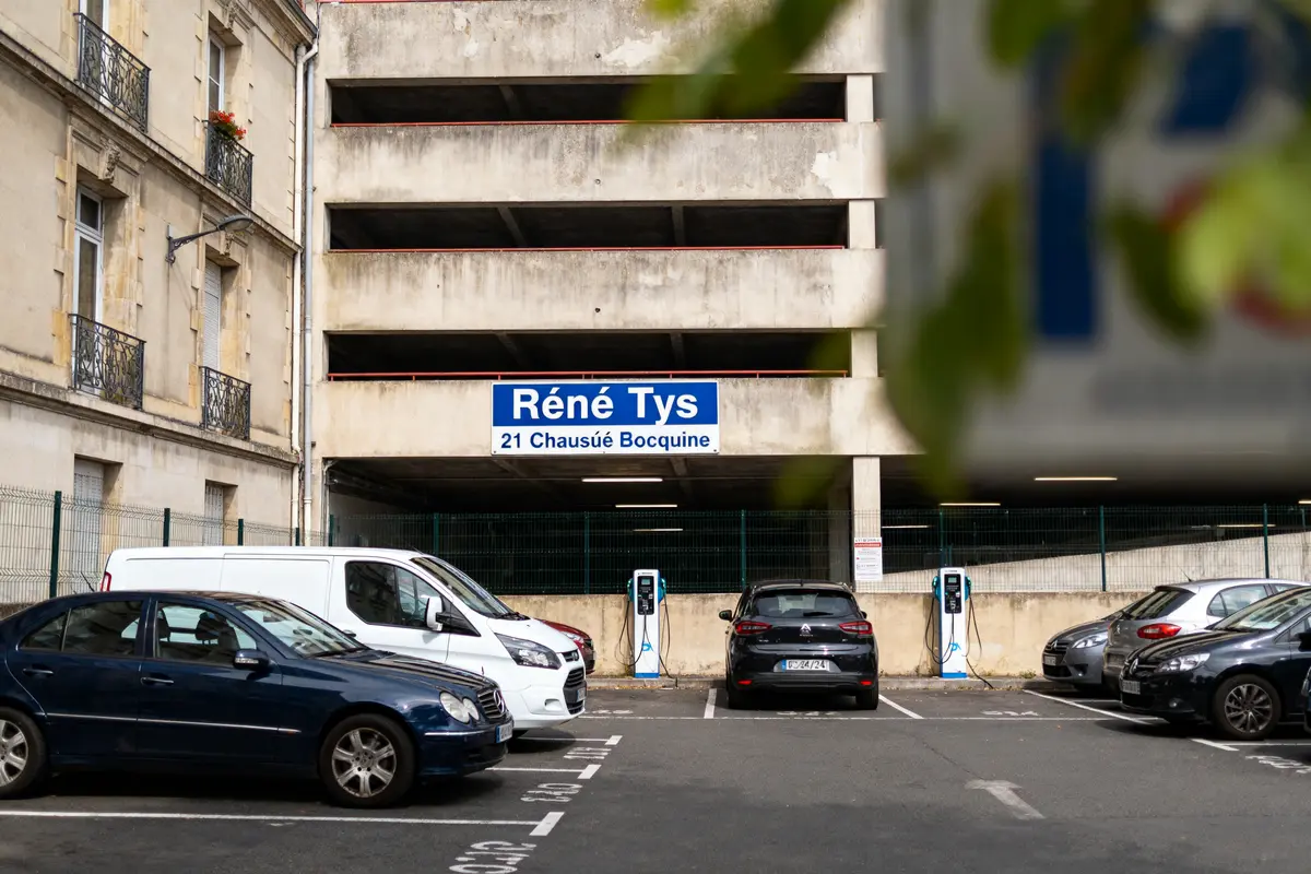 Parking avec voitures, bâtiment beige, panneau "Réné Tys", bornes de recharge. Adresse : 21 Chaussée Bocquaine.