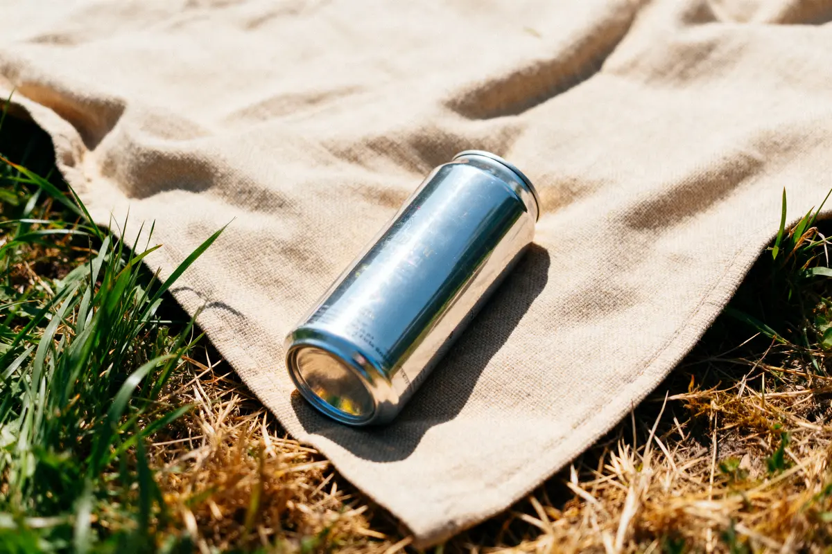 Canette métallique argentée sur une couverture beige, posée sur l'herbe ensoleillée.